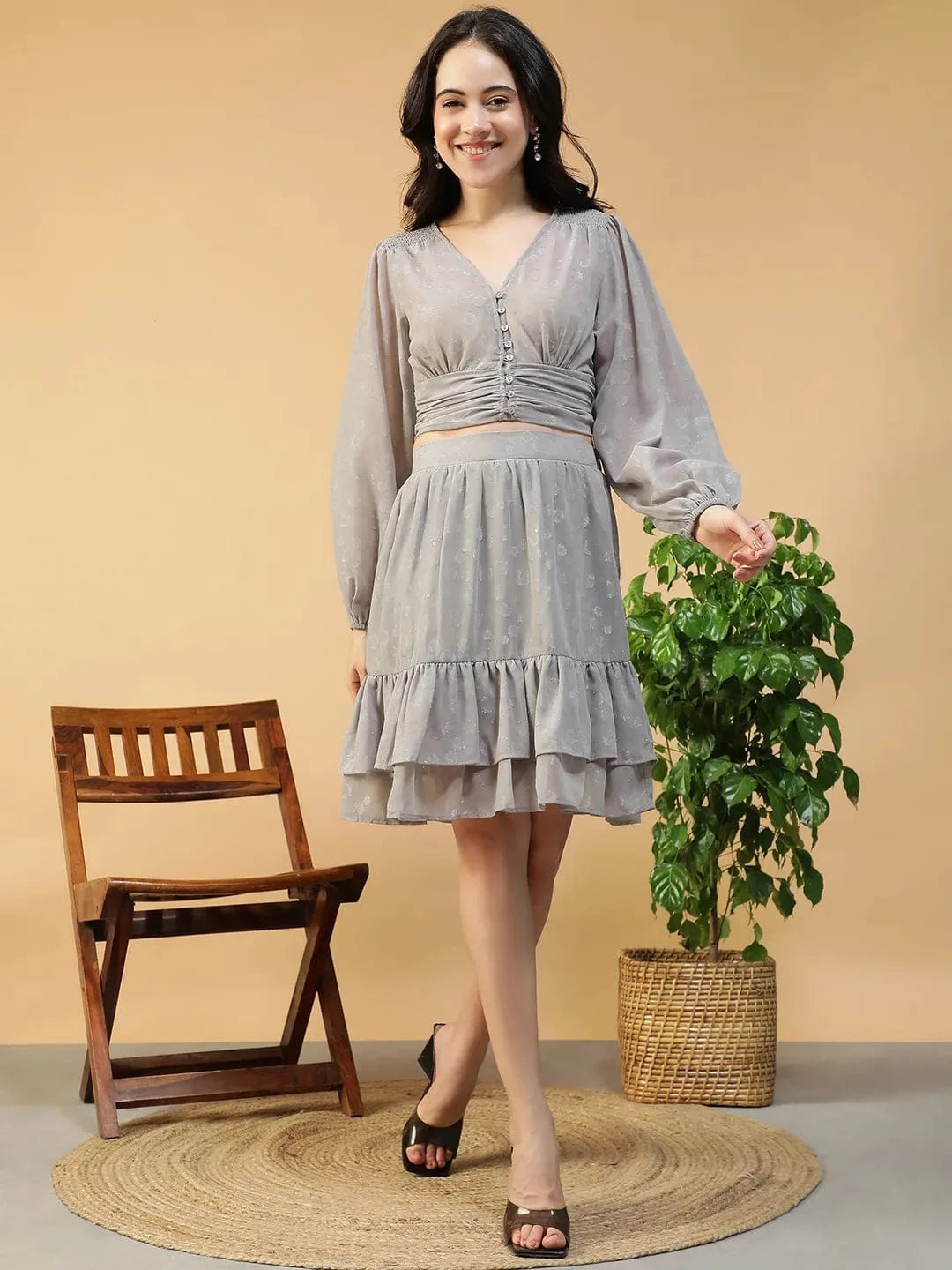 A young woman wearing a gray dress with ruffles and a gray blouse, standing in front of a wooden chair and a potted plant.