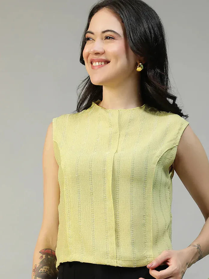 Woman wearing a light green sleeveless top and black pants against a neutral background with a plant.