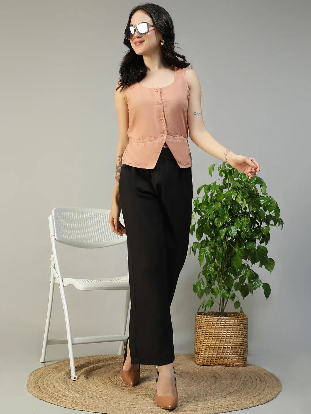 Woman in peach sleeveless top and black pants standing next to a white chair and potted plant on a gray background