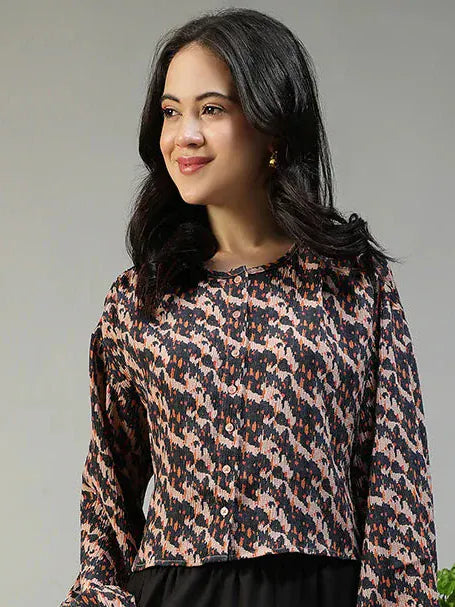 Woman in a patterned top and black pants standing next to a plant and chair on a gray background