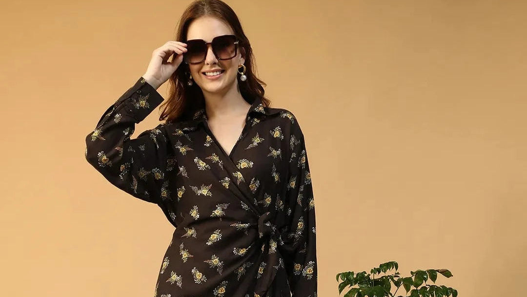 Woman in a black patterned dress standing next to a plant and brown chair against a beige wall.