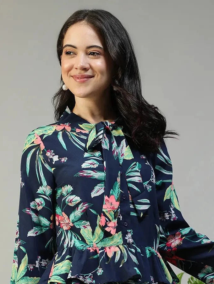 Woman wearing a navy blue floral blouse with white pants standing next to a plant on a gray background