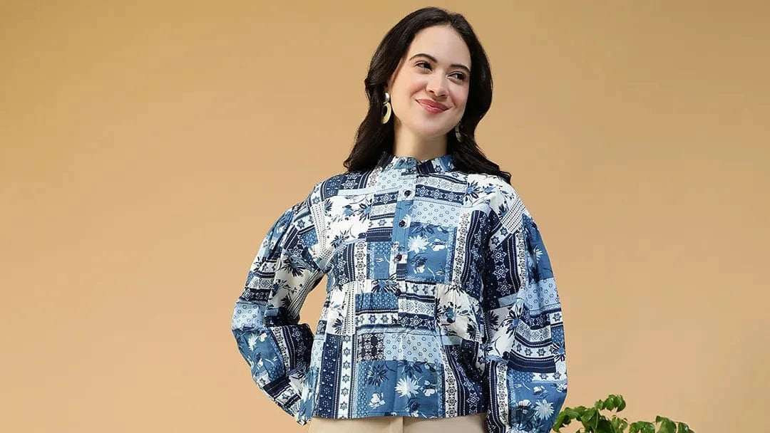 Woman wearing a blue patterned blouse and beige pants standing in a room with a wooden chair and plant.